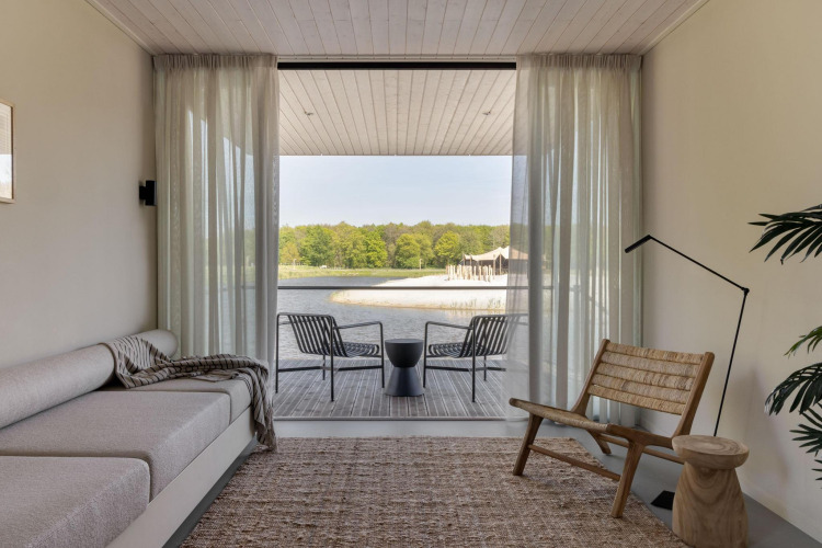 Minimalist glamping accommodation living room with sofa, wood chair and lake view through glass doors.
