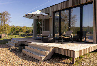 Lodge-terras uit hout met twee stoelen, parasol en glazen, grenzend aan grote ramen en zicht op de natuur.