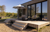 Wooden lodge deck with two chairs, umbrella, glasses and large glass doors, overlooking a peaceful nature view.