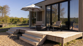 Houten terras bij een lodge met twee stoelen, parasol en glazen, naast grote glazen deuren en natuur.