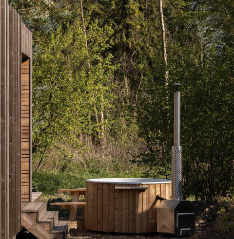 Bañera de madera con chimenea junto a una cabaña en Resort de Parel, Países Bajos, entre árboles verdes.