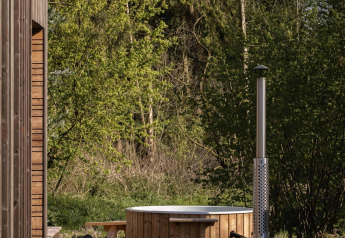 Bañera de madera con chimenea junto a una cabaña en Resort de Parel, Países Bajos, entre árboles verdes.