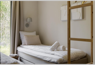 Modern lodge-kamer met eenpersoonsbed, kussens, opgerolde handdoeken en raam met uitzicht op groen.