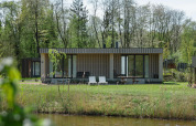 Moderne lodge met houten gevel en grote ramen, gelegen aan een vijver in een groene, bosrijke omgeving.