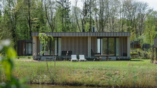 Modern lodge with wooden slats and large windows, set in green surroundings by a pond in springtime.