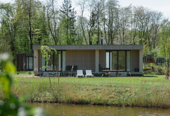 Moderne Lodge mit Holzfassade, großen Fenstern und Gartenmöbeln am See, umgeben von Bäumen im Frühling.