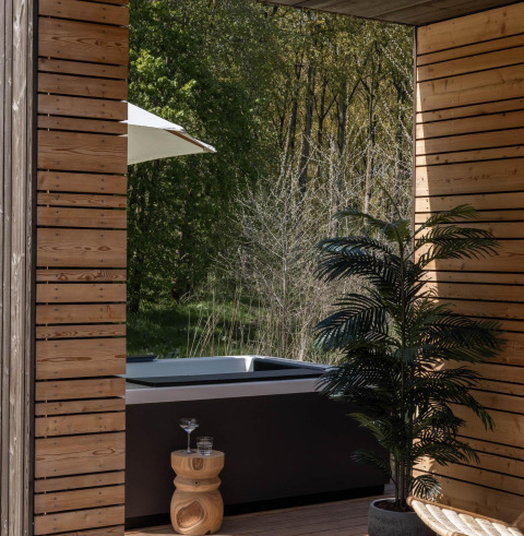 Zona exterior de lodge con paneles de madera, jacuzzi, plantas y vista a un bosque verde y tranquilo.