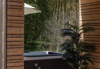Zona exterior de lodge con paneles de madera, jacuzzi, plantas y vista a un bosque verde y tranquilo.