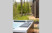 Jacuzzi exterior sobre una tarima de madera junto a una cabaña, rodeado de árboles y naturaleza verde.