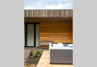 Vista exterior en una cabaña con fachada de madera, terraza, banco y jacuzzi bajo un cielo parcialmente nublado.