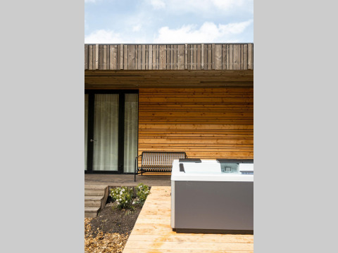 Vista exterior en una cabaña con fachada de madera, terraza, banco y jacuzzi bajo un cielo parcialmente nublado.