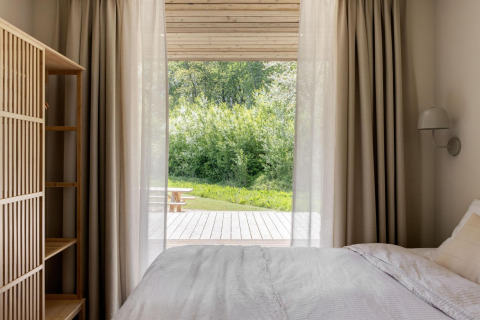 Dormitorio acogedor en lodge con cortinas claras, cama blanca y vistas a terraza y naturaleza verde.