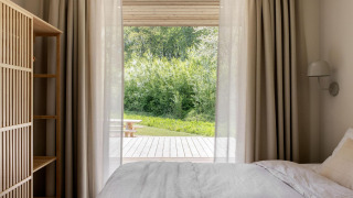 Gezellige slaapkamer in een lodge met lichte gordijnen, bed en uitzicht op terras en groene natuur.