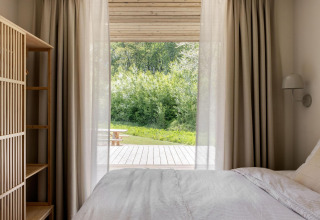 Dormitorio acogedor en lodge con cortinas claras, cama blanca y vistas a terraza y naturaleza verde.