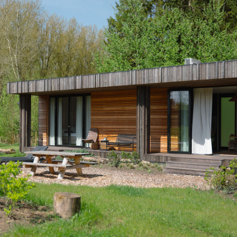 Photo extérieure d’un lodge moderne en bois avec terrasse, bancs de jardin, table de pique-nique et verdure.