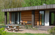 Photo extérieure d’un lodge moderne en bois avec terrasse, bancs de jardin, table de pique-nique et verdure.