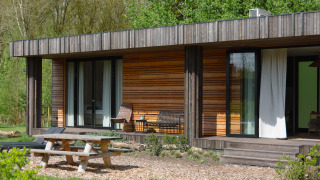 Außenansicht einer modernen Holzhütte mit Terrasse, Sitzgelegenheiten, Picknicktisch und Naturumgebung.