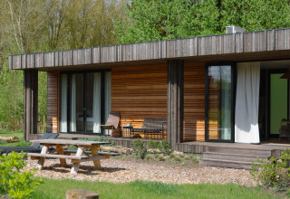 Außenansicht einer modernen Holzhütte mit Terrasse, Sitzgelegenheiten, Picknicktisch und Naturumgebung.