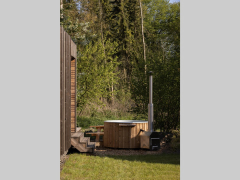 Bañera de hidromasaje de madera al aire libre junto a una cabaña, rodeada de naturaleza y bosque verde.