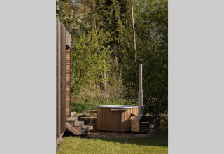Houten buitenbubbelbad naast een lodge, omgeven door groene bomen en gras op een zonnige lentedag.