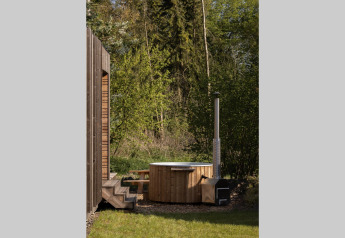 Bain à remous extérieur en bois près d’un lodge, entouré de verdure et de forêt sous un ciel ensoleillé.