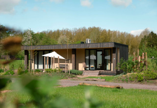 Un lodge moderne dans un cadre verdoyant, avec terrasse, chaises et parasol, sous une belle journée ensoleillée.