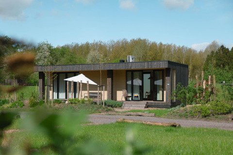 Un lodge moderne dans un cadre verdoyant, avec terrasse, chaises et parasol, sous une belle journée ensoleillée.