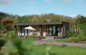 A modern lodge cabin set in lush greenery, featuring a terrace with seating and a sun umbrella on a sunny day.