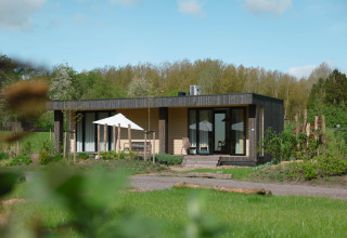 Et moderne lodgehus i grønne omgivelser med terrasse, udendørs siddepladser og parasol på en solrig dag.