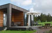 Villa moderne en bois avec terrasse, chaises longues et parasol au Resort de Parel, entourée de verdure.