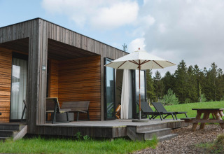 Villa moderne en bois avec terrasse, chaises longues et parasol au Resort de Parel, entourée de verdure.