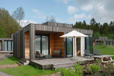Moderna cabaña de madera con terraza, tumbonas y sombrilla en un campo verde rodeado de árboles y otras cabañas.