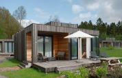 Moderne houten lodge met terras, ligstoelen en parasol op een grasveld tussen bomen en andere huisjes.