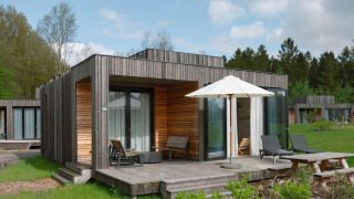 Moderne houten lodge met terras, ligstoelen en parasol op een grasveld tussen bomen en andere huisjes.