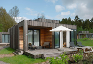 Moderne houten lodge met terras, ligbedden en parasol op een groene weide, omringd door bomen en huisjes.