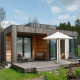 Moderne træhytte med terrasse og liggestole på en grøn eng ved siden af andre hytter, omgivet af træer.