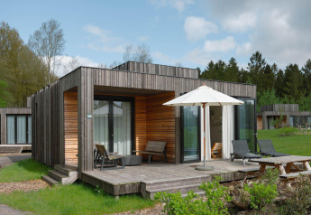 Moderna cabaña de madera con terraza, tumbonas y sombrilla en un campo verde rodeado de árboles y otras cabañas.