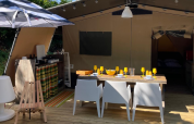 Safari Base Lodge en Camping Gajole, Italia, con tienda abierta, mesa para comer, cocina y sombrilla exterior.