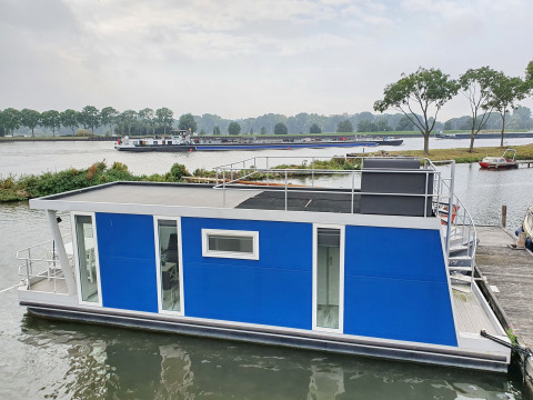 Modern blue houseboat docked at Marina Parcs Amsterdam, Netherlands, with water and trees behind.