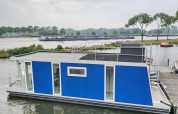 Blå husbåd ved en havn i Amsterdam, med terrasse ovenpå og grønne træer i baggrunden.