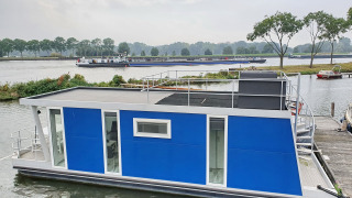 Blå husbåd ved en havn i Amsterdam, med terrasse ovenpå og grønne træer i baggrunden.