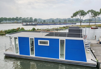 Blå husbåd ved en havn i Amsterdam, med terrasse ovenpå og grønne træer i baggrunden.
