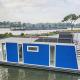 Modern blue houseboat docked at Marina Parcs Amsterdam, Netherlands, with water and trees behind.