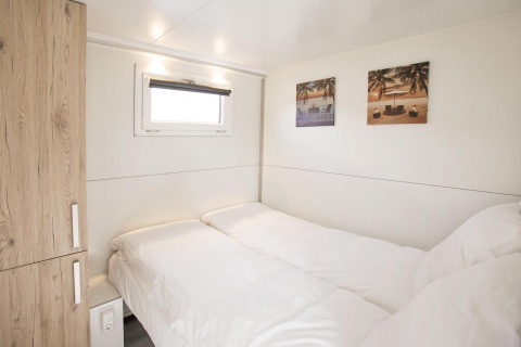 Slaapkamer op een woonboot met twee enkele bedden, houten kast, klein raam en strandfoto’s aan de muur.