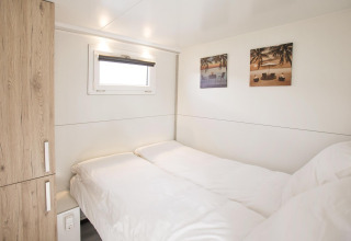 Slaapkamer op een woonboot met twee enkele bedden, houten kast, klein raam en strandfoto’s aan de muur.