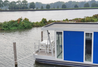 Blue houseboat named Havenlodge+ moored at Marina Parcs Amsterdam, with river and greenery in background.
