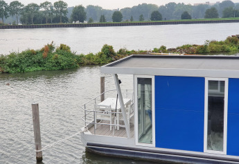 Casa flotante azul Havenlodge+ amarrada en Marina Parcs Amsterdam, junto al río y vegetación al fondo.