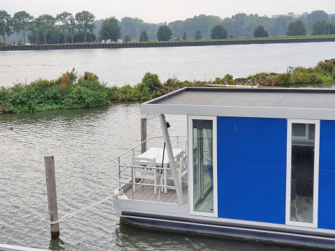 Blue houseboat named Havenlodge+ moored at Marina Parcs Amsterdam, with river and greenery in background.