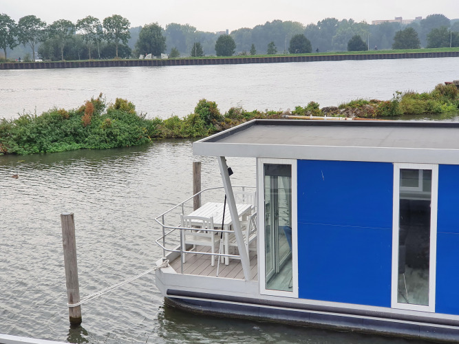 Casa flotante azul Havenlodge+ amarrada en Marina Parcs Amsterdam, junto al río y vegetación al fondo.