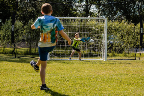 Twee kinderen spelen voetbal bij een doel op het gras bij een safaritent, omgeven door bomen.
